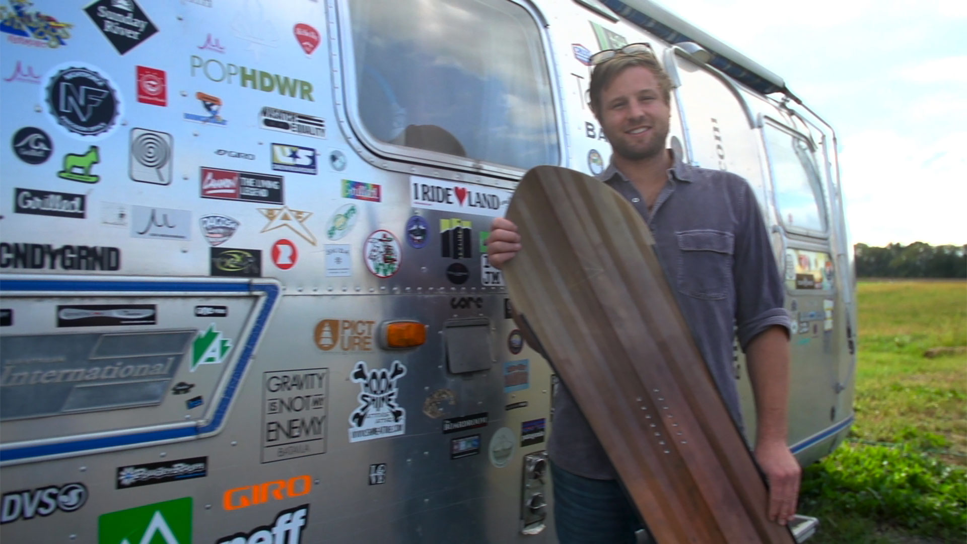 Man holding a snowboard standing in front of an airstream trailer covered in decals