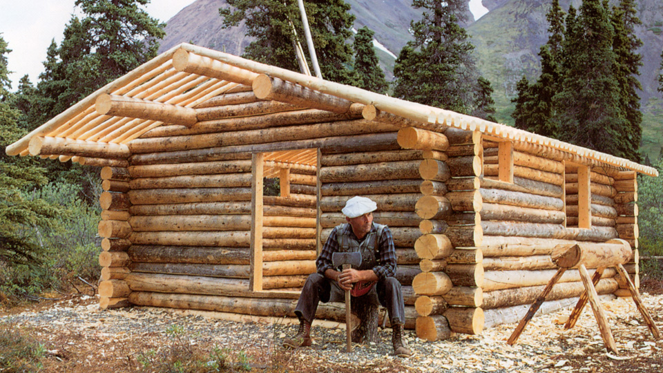 Dick Proenneke sitting in front of his log cabin