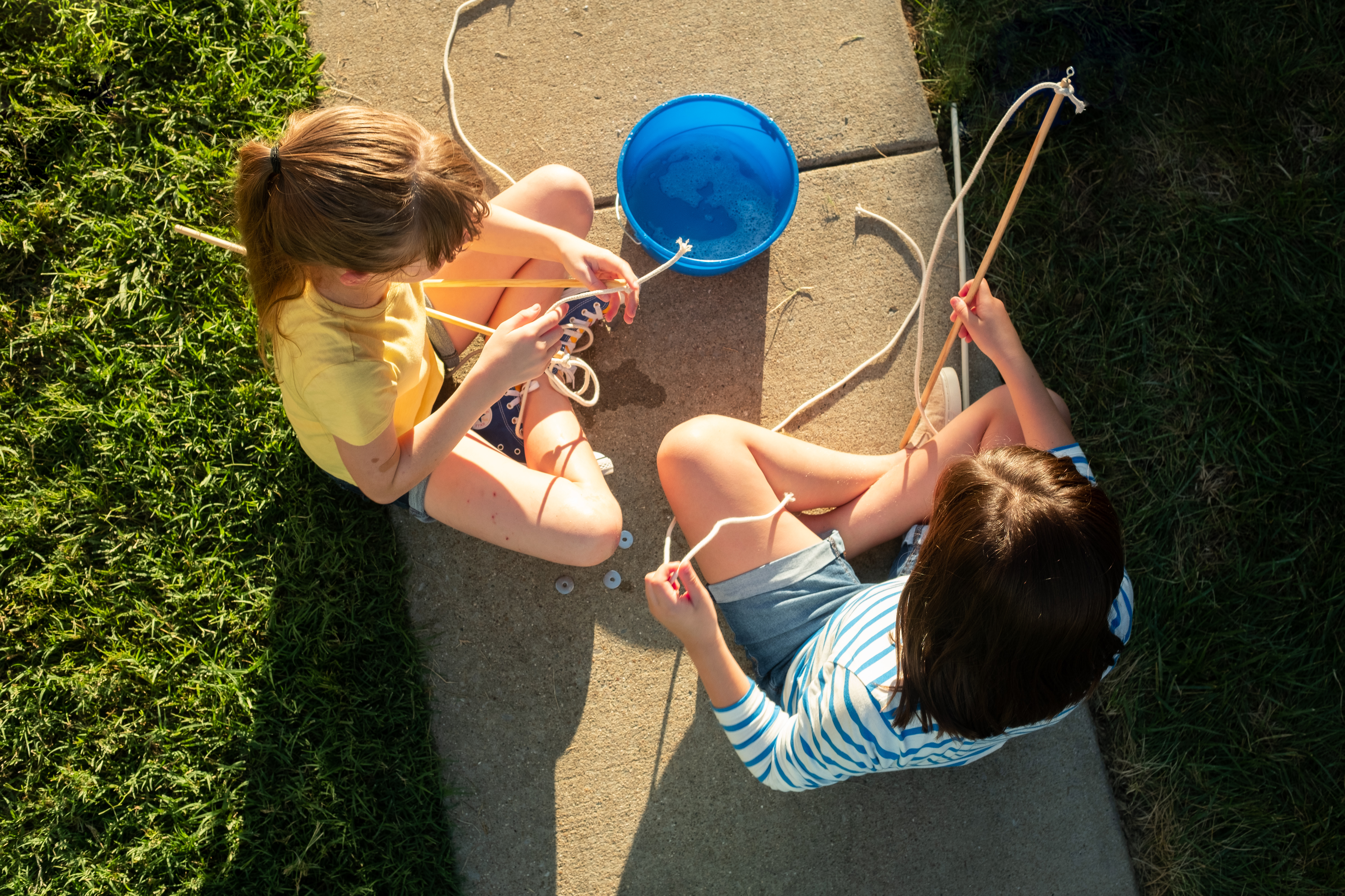 A girl in a yellow shirt and a girl in a blue and white striped shirt sit on a sidewalk next to a blue bucket filled with soapy water.