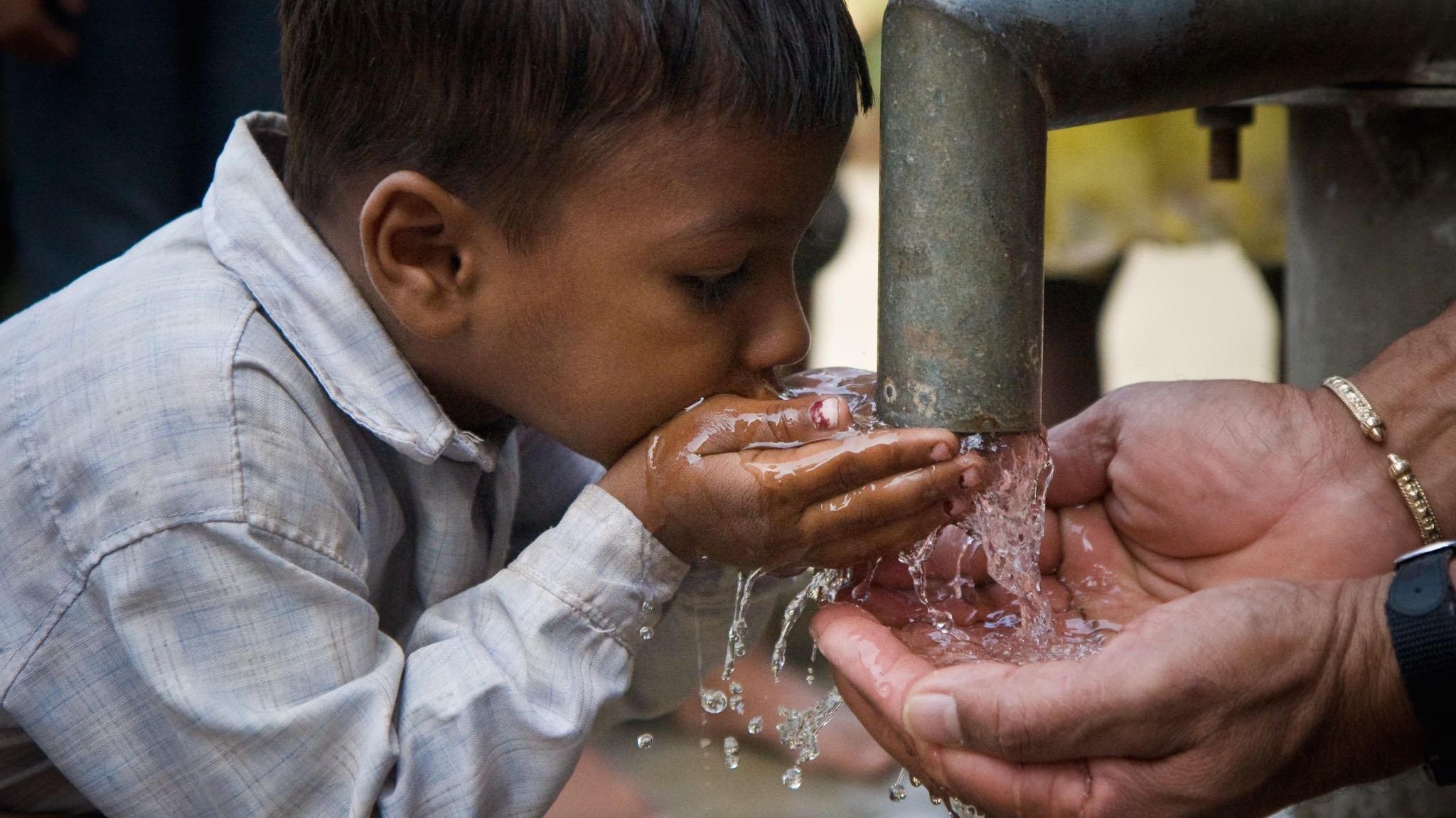 A boy getting water to drink out of a pipe with adult hands nearby.