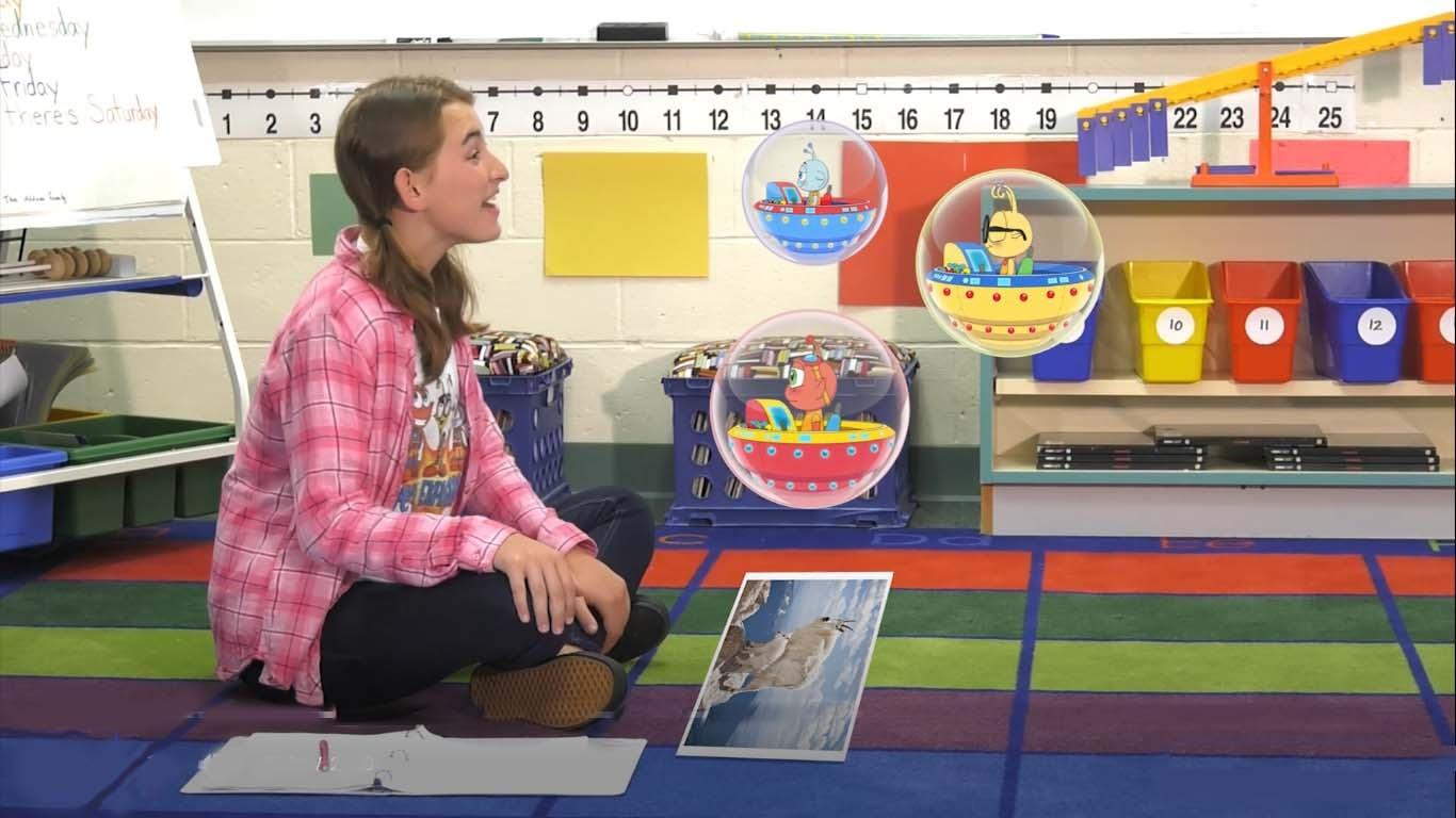 A girl sits on the floor and talks to three cartoon characters in spaceships.