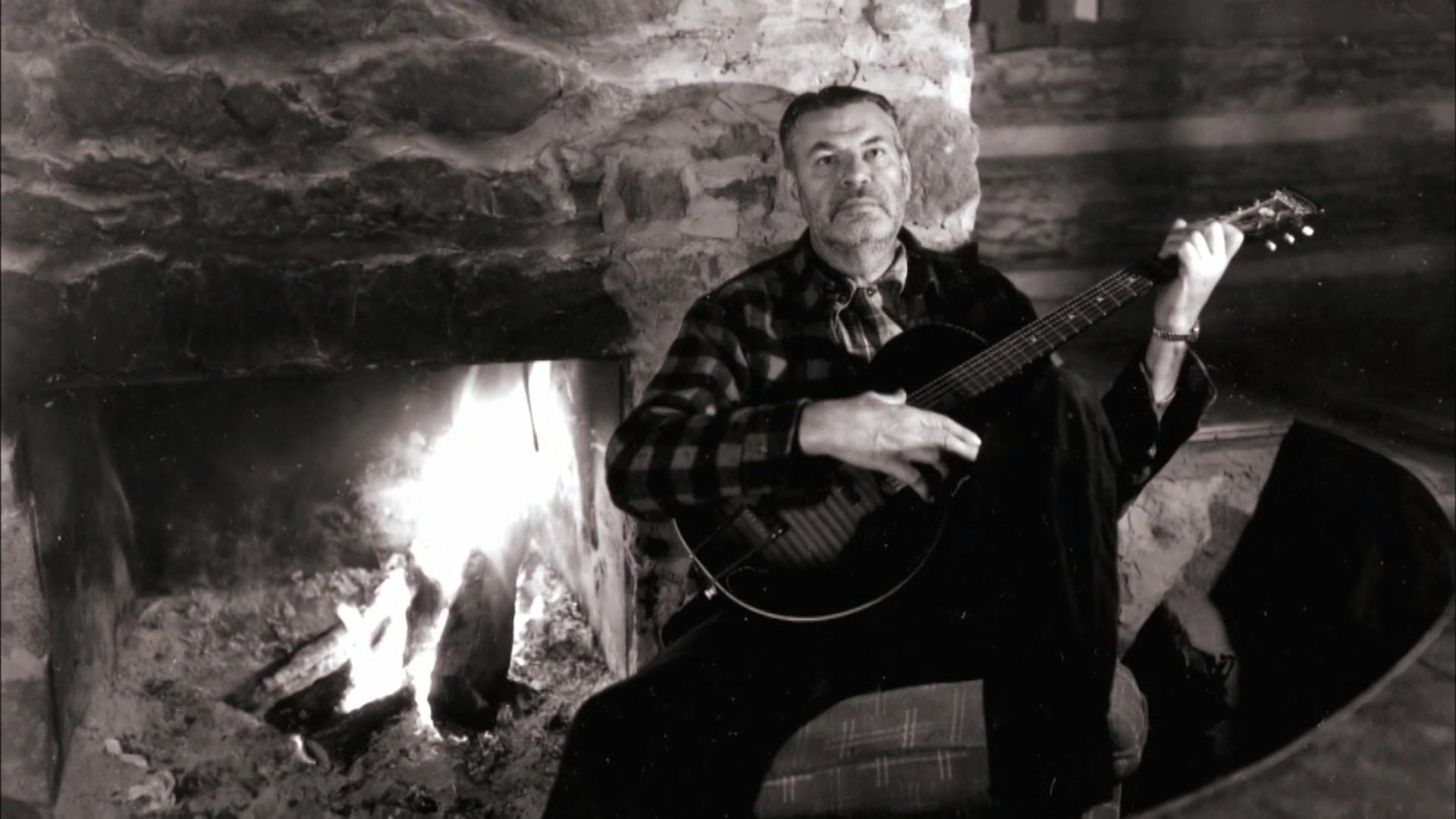A man with a guitar sits in front of a fireplace.