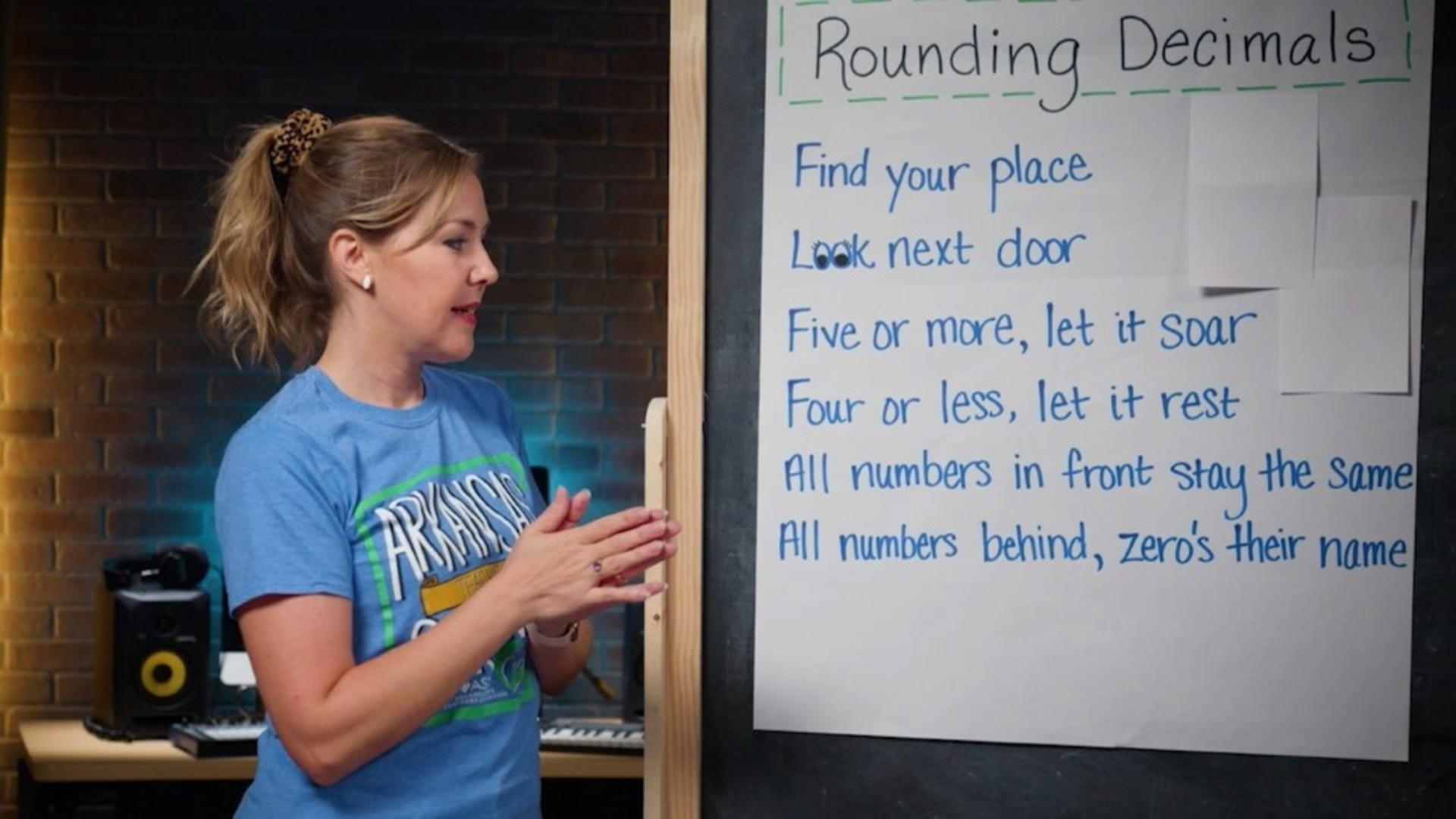 A woman in a blue shirt stands in front of a chalkboard with a giant stickie note on it.