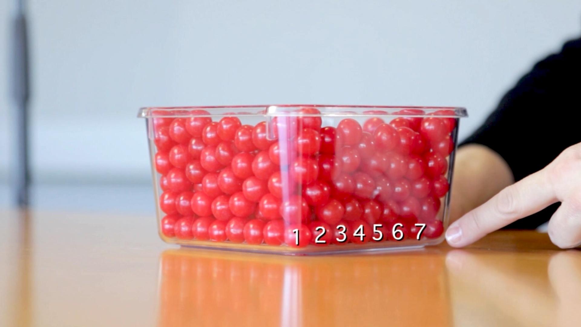 A box of red candy with a person pointing to pieces to estimate volume.