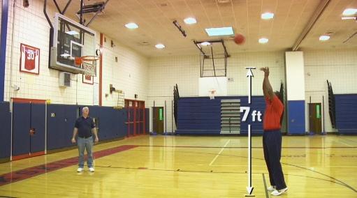 Two men stand in a basketball court where one shoots a ball at a hoop.