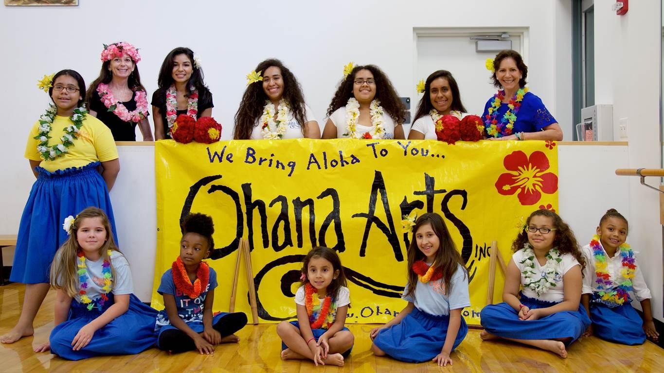 A group of children in Hawaiian dance wear sit and stand around a banner.