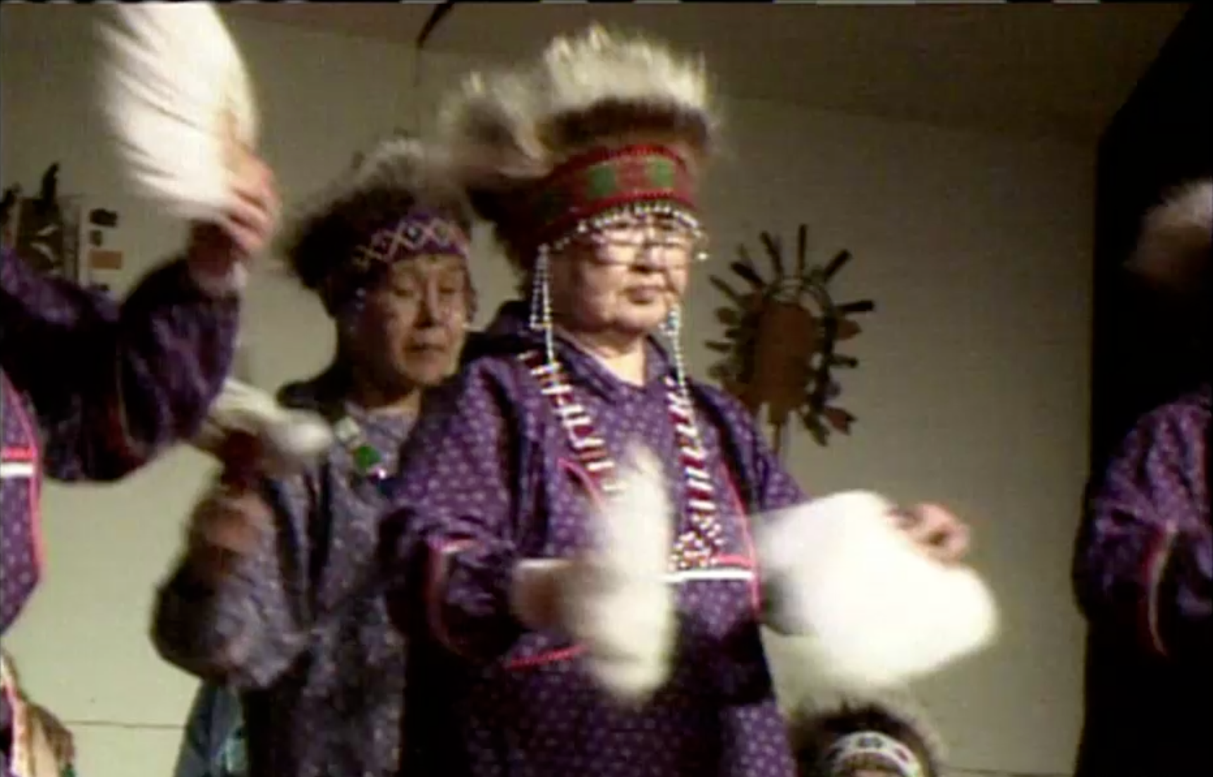 Three dancers in regalia hold objects as they dance.