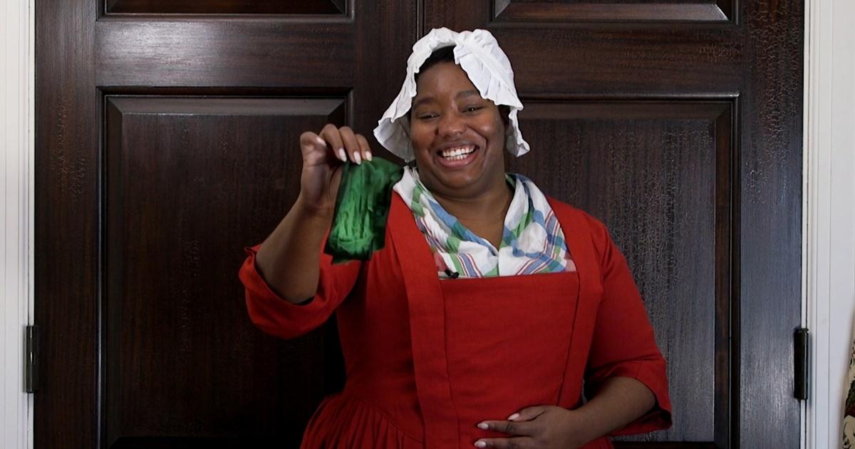 A Black woman in historical dress holds an object up.
