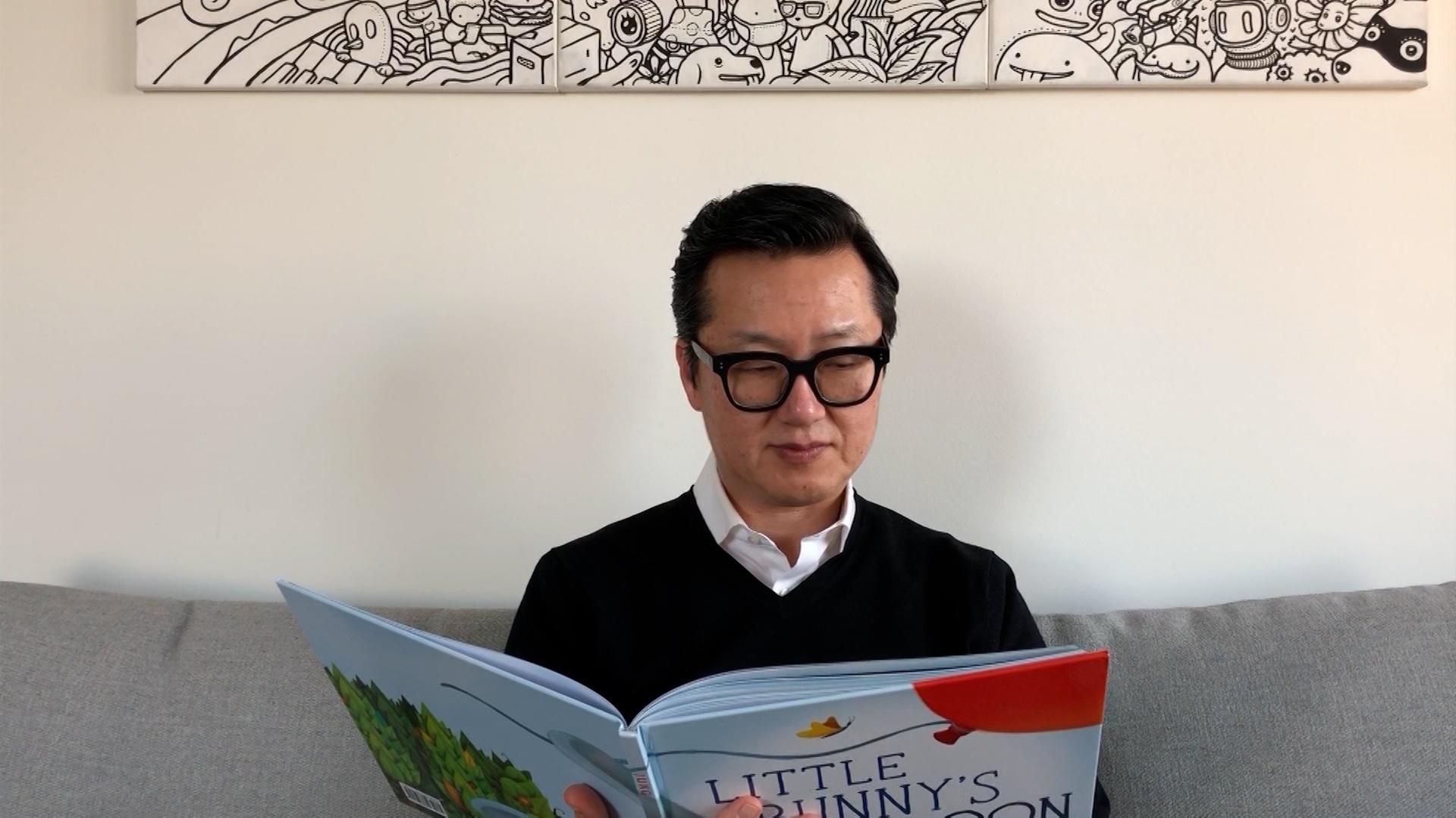 An Asian man with glasses reads a children's book.