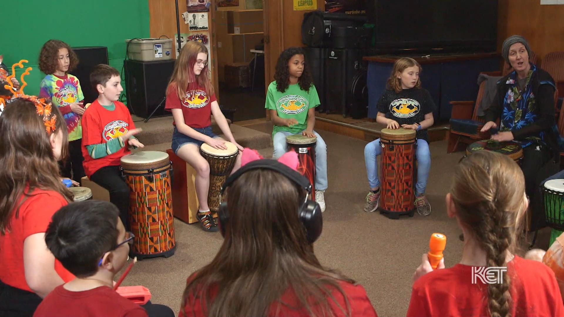 A group of children with African drums and other instruments sit in a circle playing.