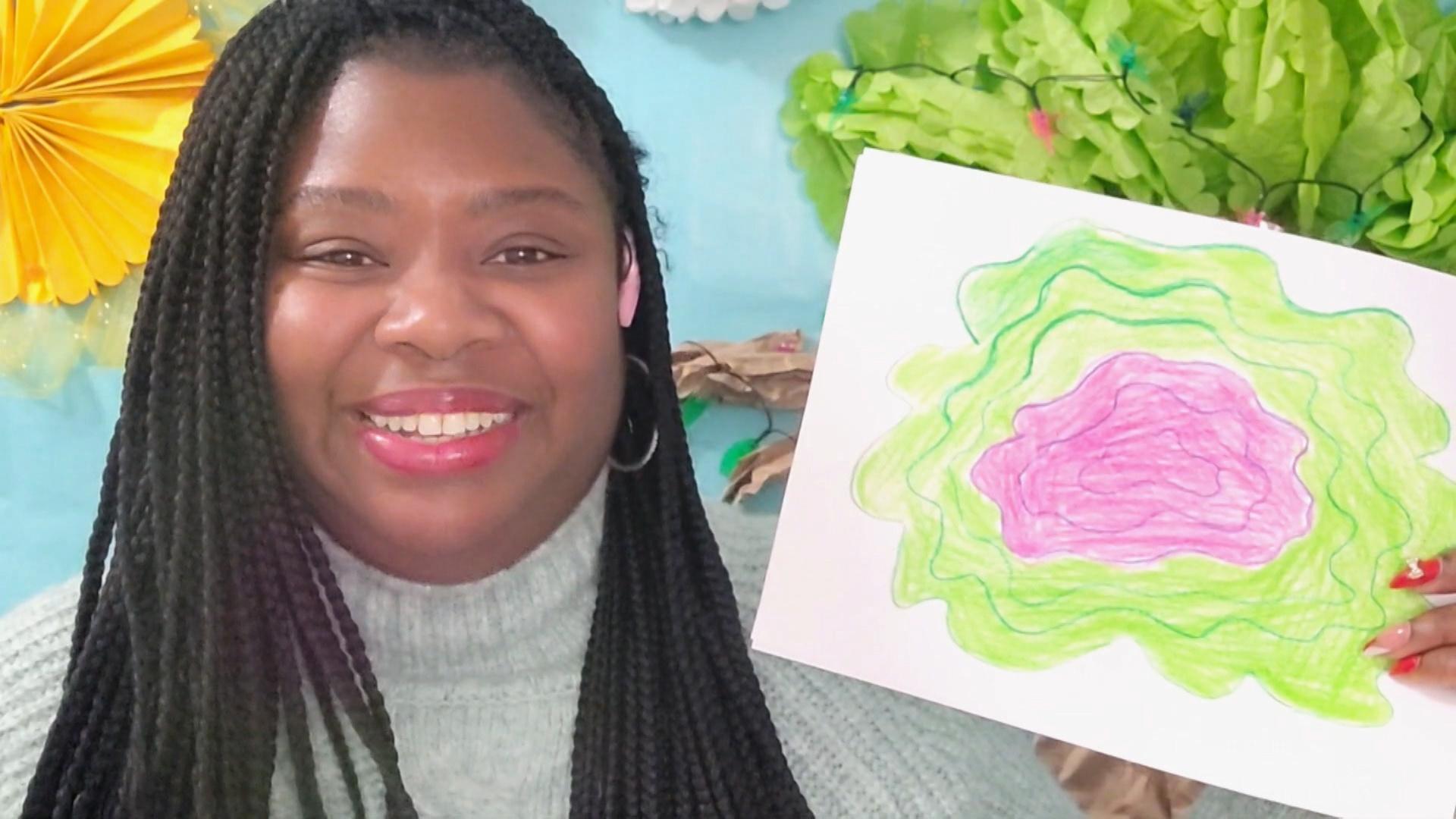 A Black woman in a grey sweater holds up a drawing with green and pink squiggles.