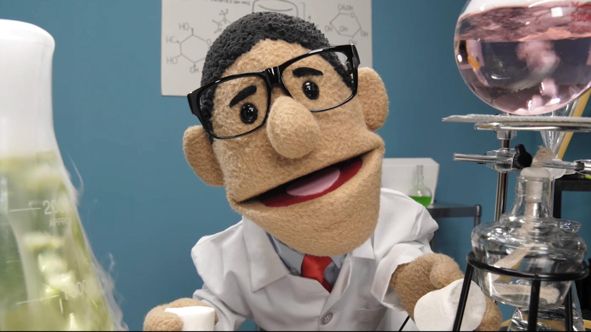 A puppet with glasses in a white lab coat stands behind laboratory equipment.