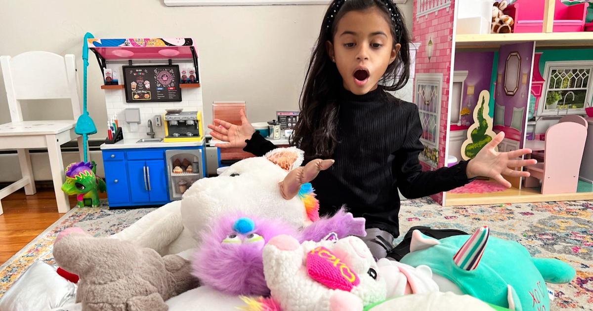 A young girl sits behind a pile of stuffed animals with a shocked look on her face.