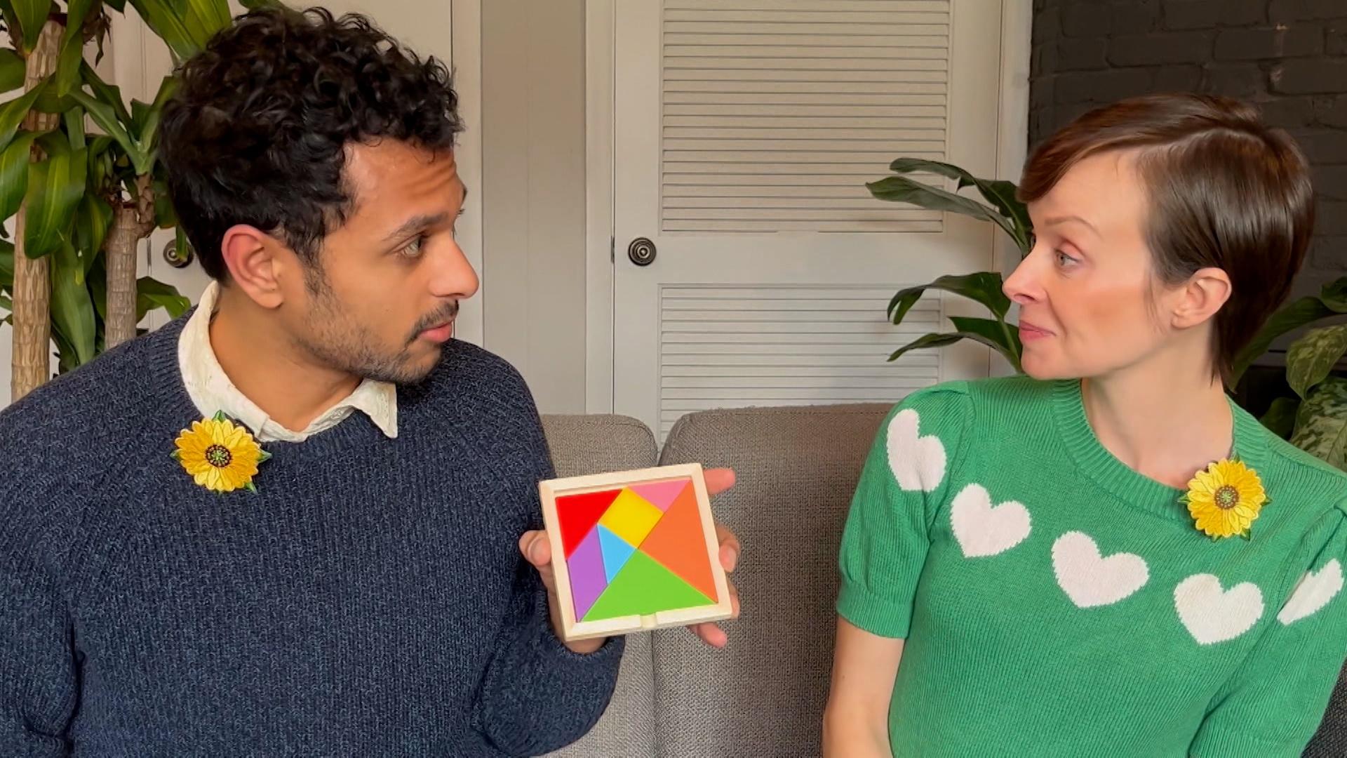 A man in a blue sweater and a woman in a green sweater sit on a tan couch, facing each other, and holding a tangram set between them.