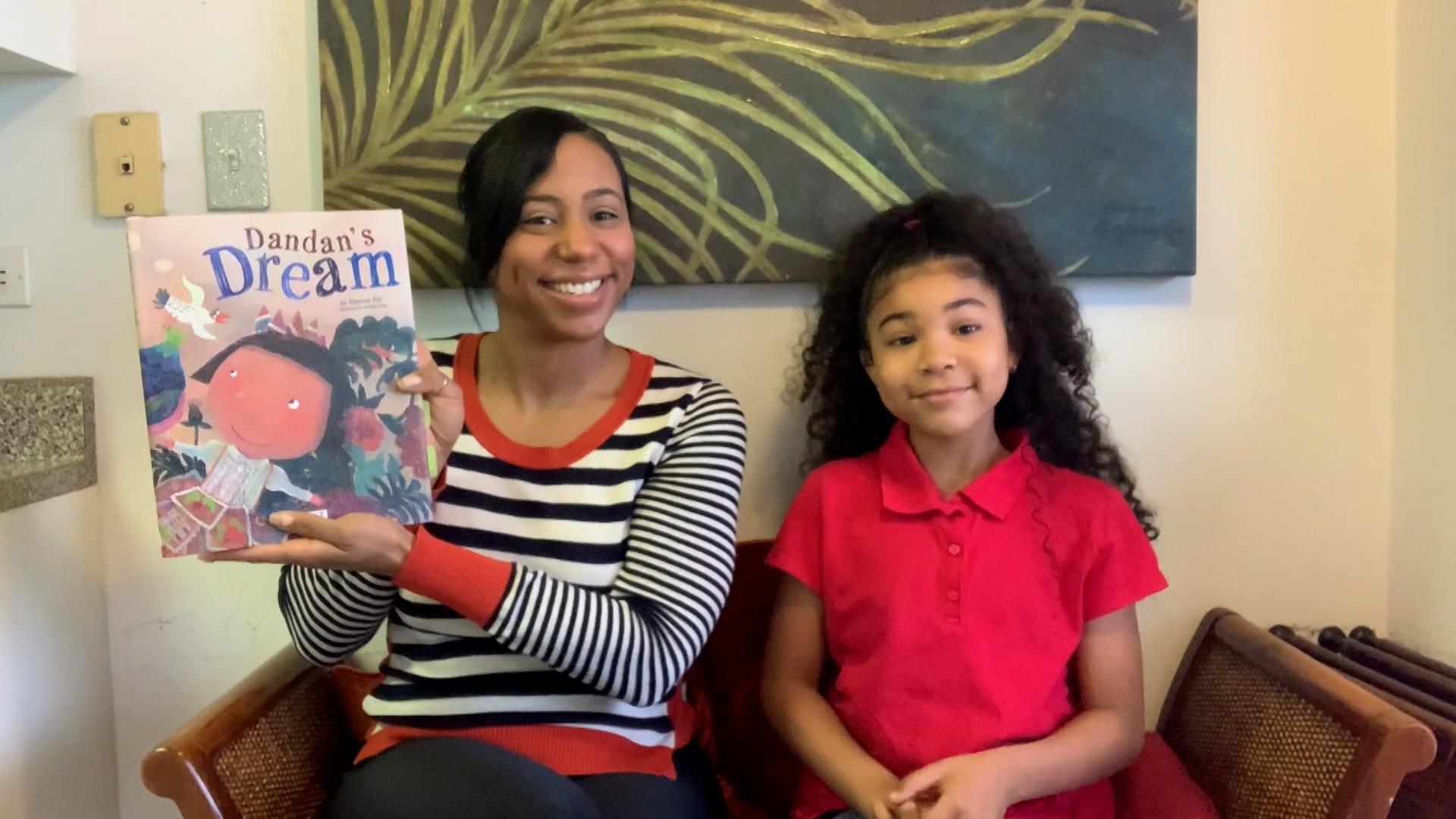 A Black woman on in a striped shirt and a Black girl in a red shirt sit on a couch and hold up a children's book.