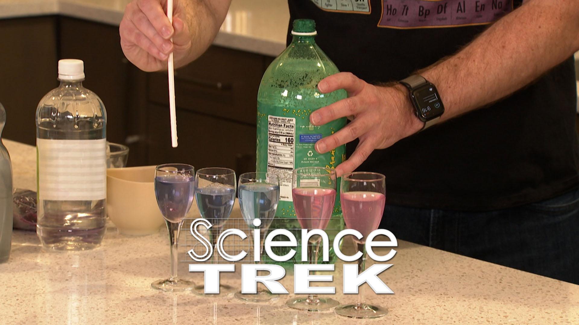 Several glasses with different color liquids stand on a counter in front of a bottle of soda.