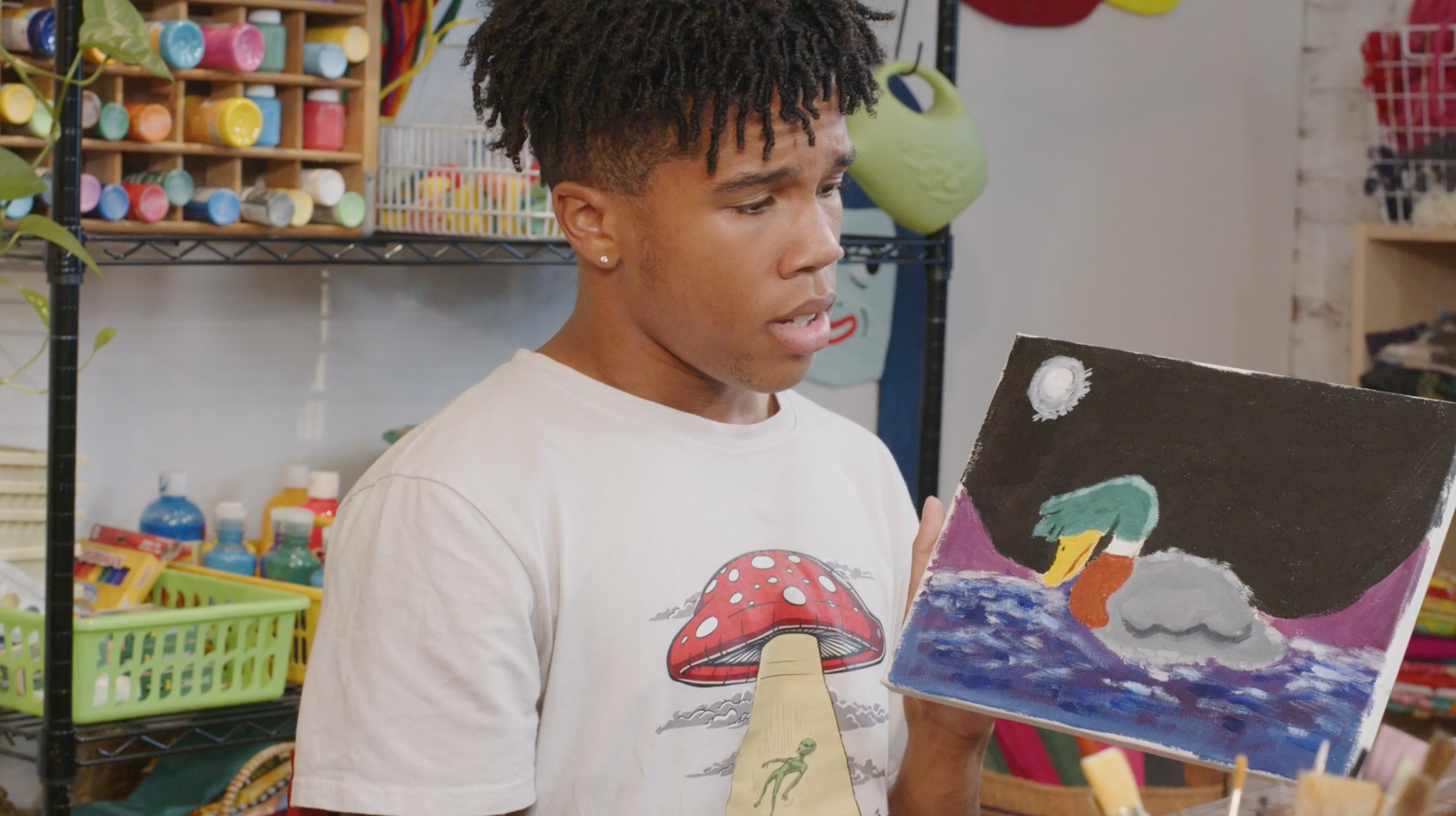 A Black man in a white t-shirt holds up a painting of a Mallard duck.