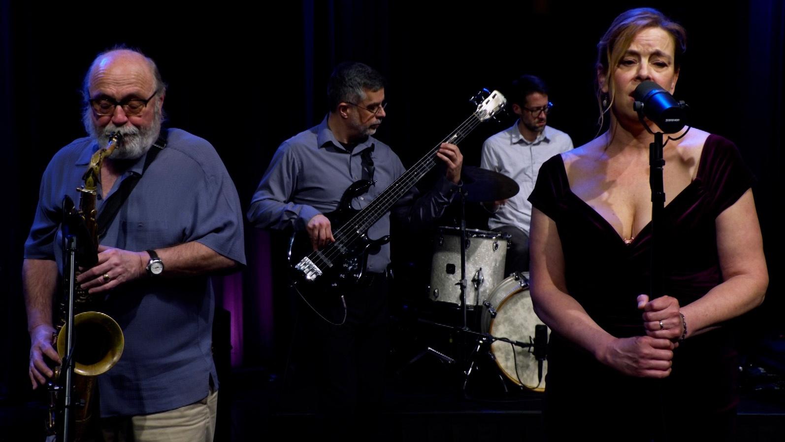 Four middle age men playing musical instruments along with woman singing into a microphone.