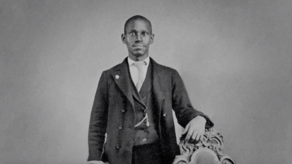 Old timey grainy portrait of a black man in a black suit standing while resting his arm on the back of a fancy chair.