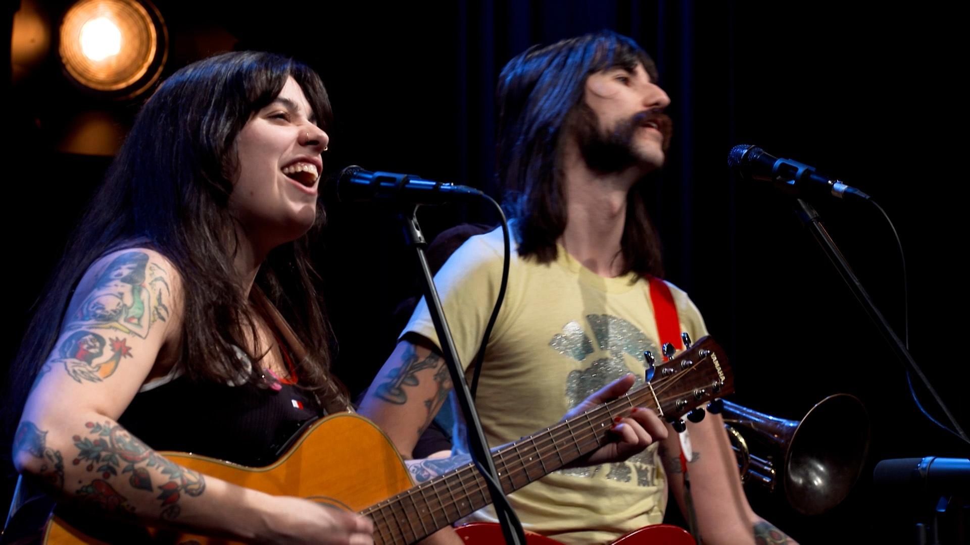 Woman playing guitar and singing into a microphone next to a man playing guitar.