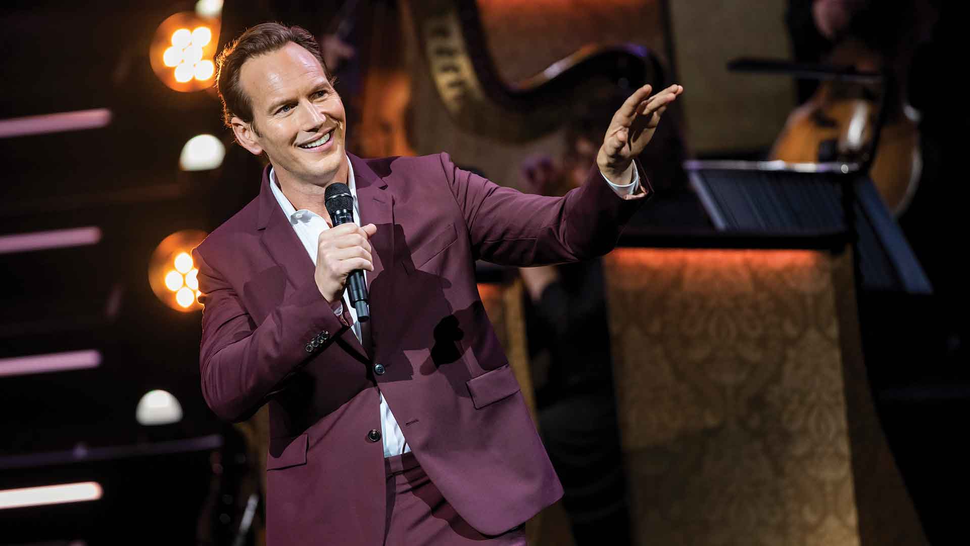 Patrick Wilson dressed in a purple suit performing on stage with a microphone in hand