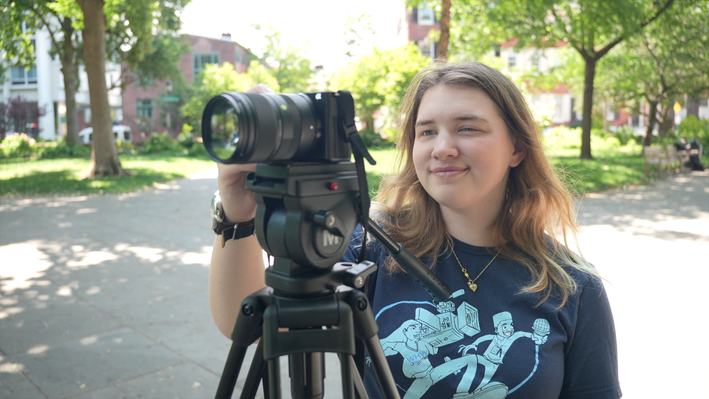 College-age girl setting up a camera with a large lens on a tripod in an outdoor campus setting.