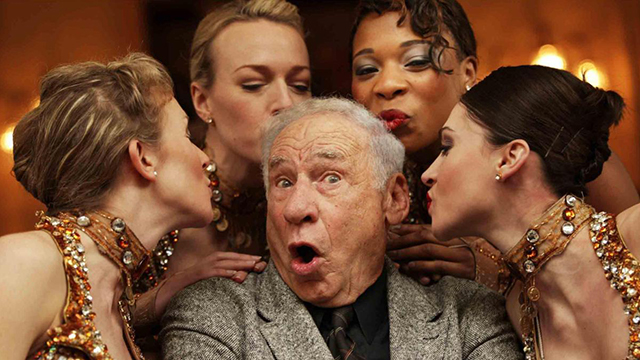 Mel Brooks with a surprised look while being surrounded by four young women.