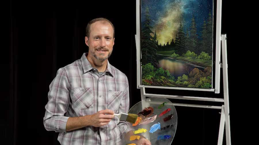 Nicolas Hankins smiling while holding a paint brush and pallet, standing in front of a painting of a night time wooded landscape on an easel.