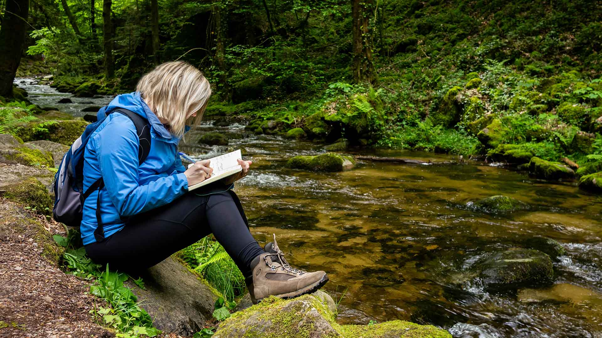 Woman sitting on the edge of a creek in a wooded area writing in a notebook.