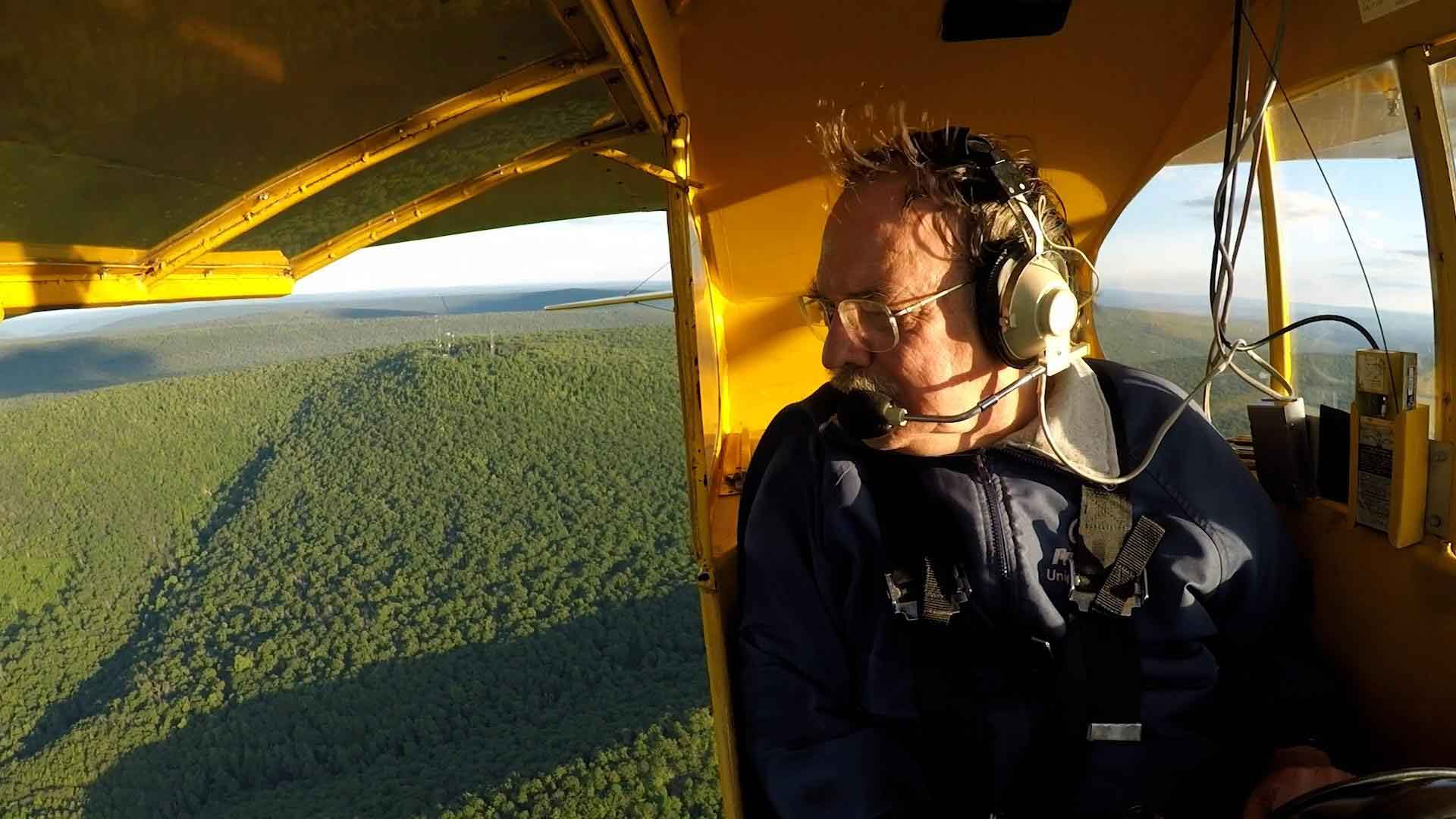 Man in a small yellow aircraft looking over shoulder at the wooded landscape below.