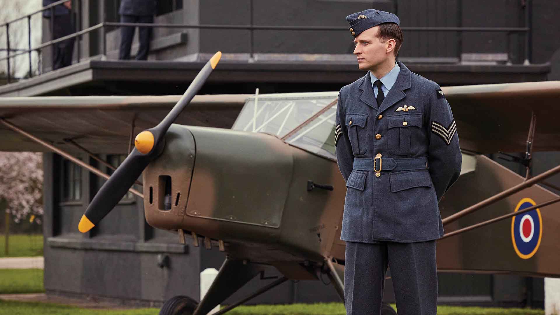 Man in military uniform standing next to a WWII era small fighter plane.