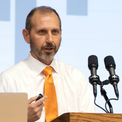 Man with beard dressed in white shirt and orange tie speaking at a podium.