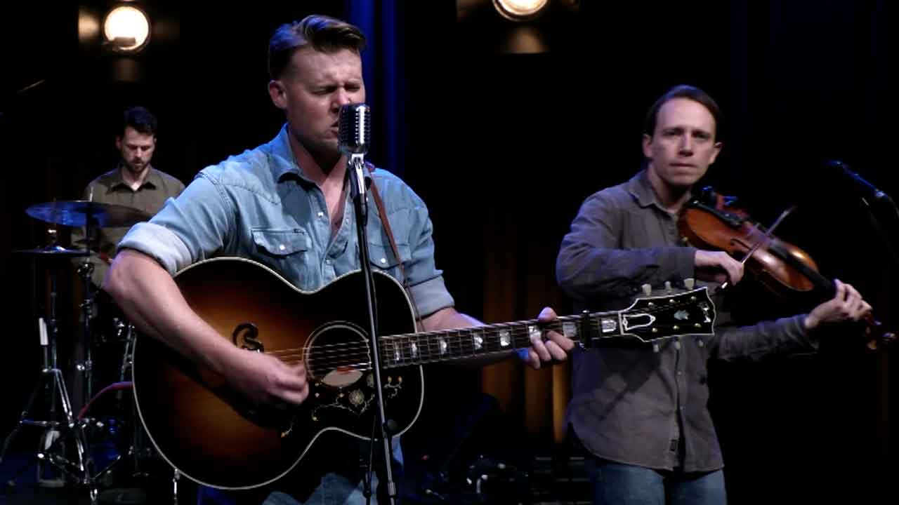 Man playing an acousting guitar and singing into a microphone with man playing violin and another man playing drums in the background.