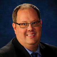 Man wearing glasses in a black suit coat with a blue shirt.