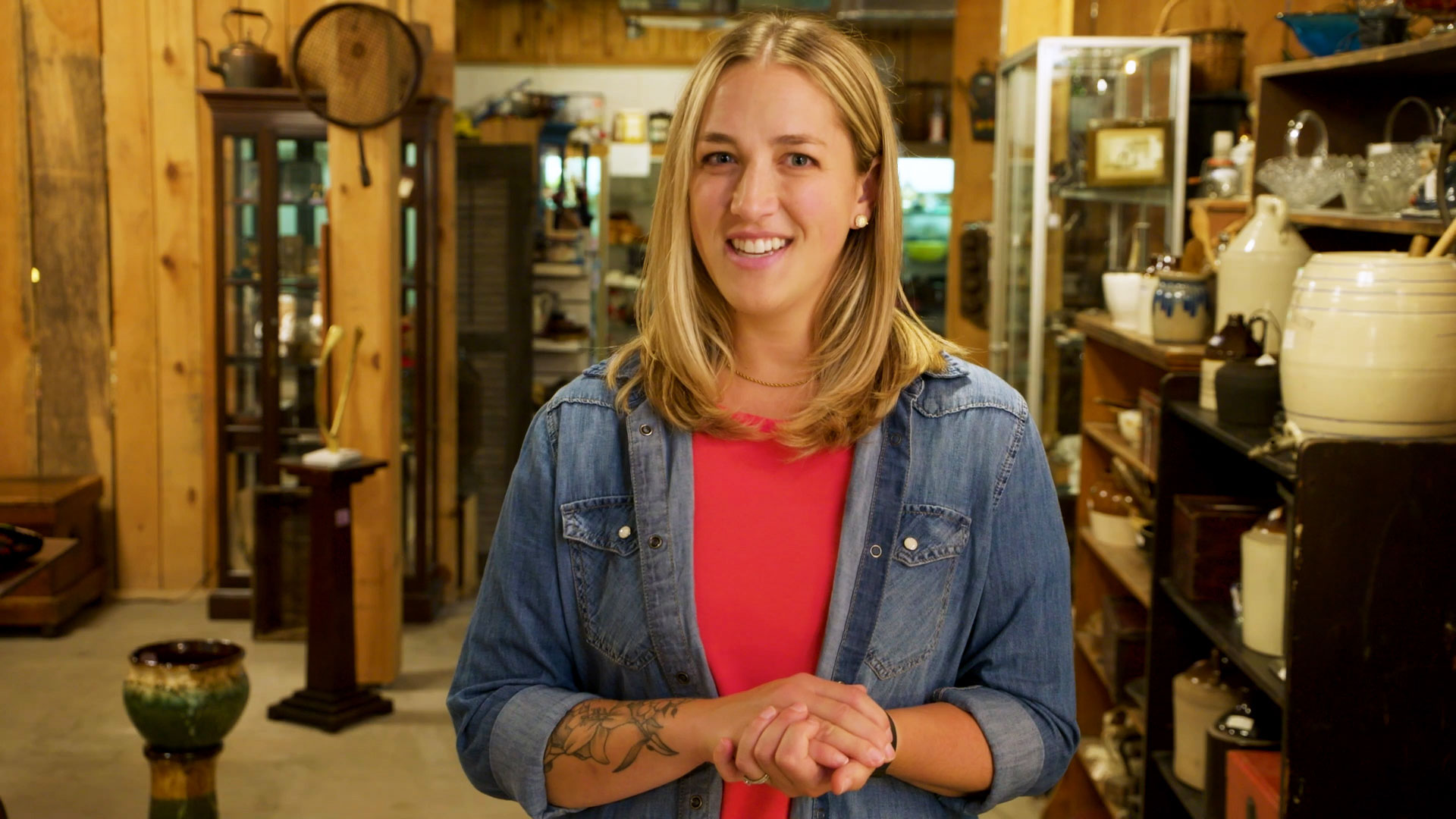 Woman with long blonde hair dressed in a denim long sleeve shirt with red undershirt, standing among various antique items on shelves.