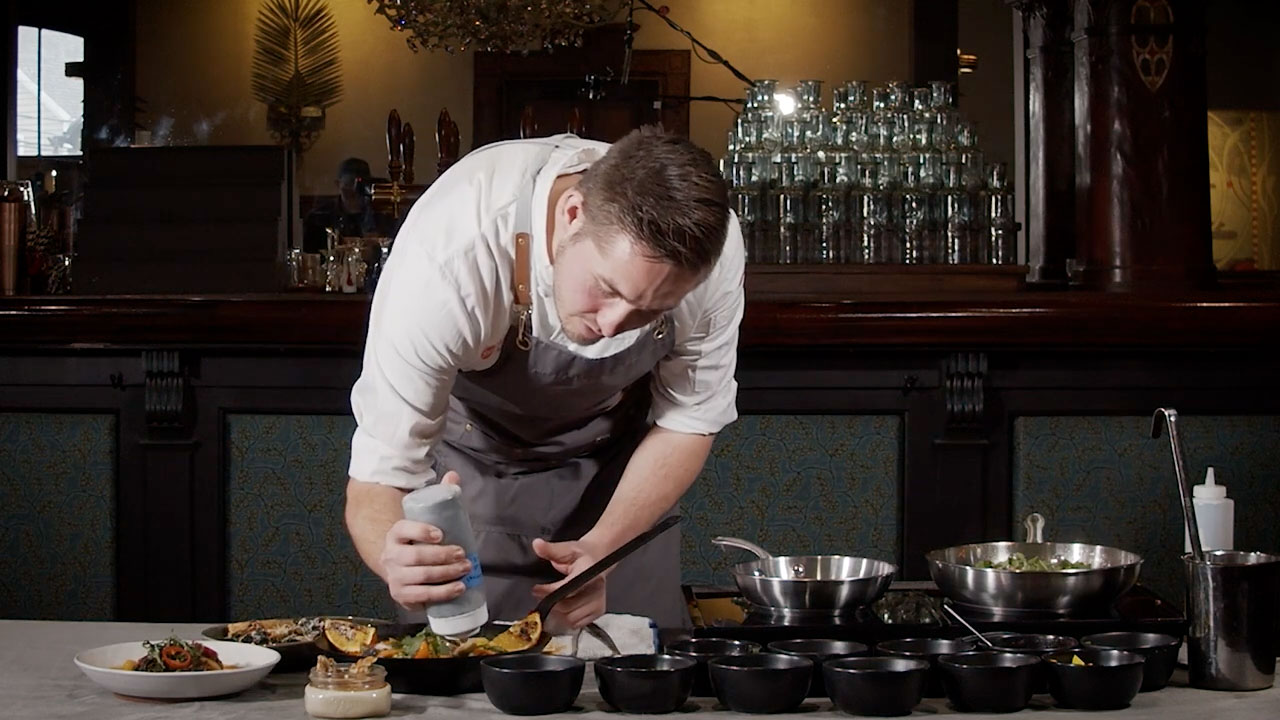 Man in a white shirt and gray apron bending over to plate food.