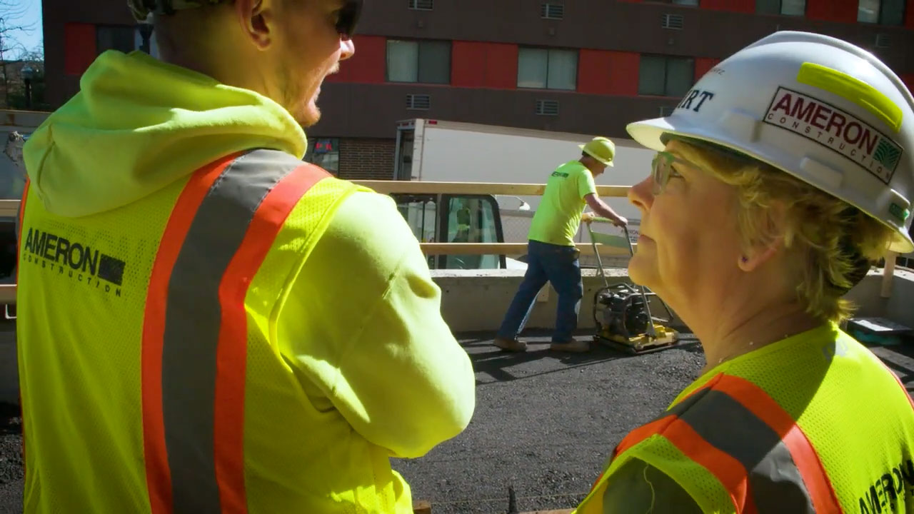 Woman in a hard hat and yellow vest talking to a man while a man operates a compactor in the background.