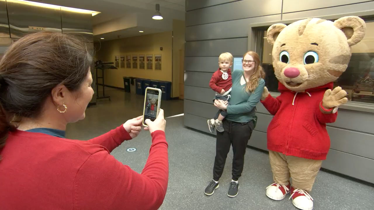 Woman in red sweater using smartphone to take a photo of a family next to a Daniel Tiger costumed character.