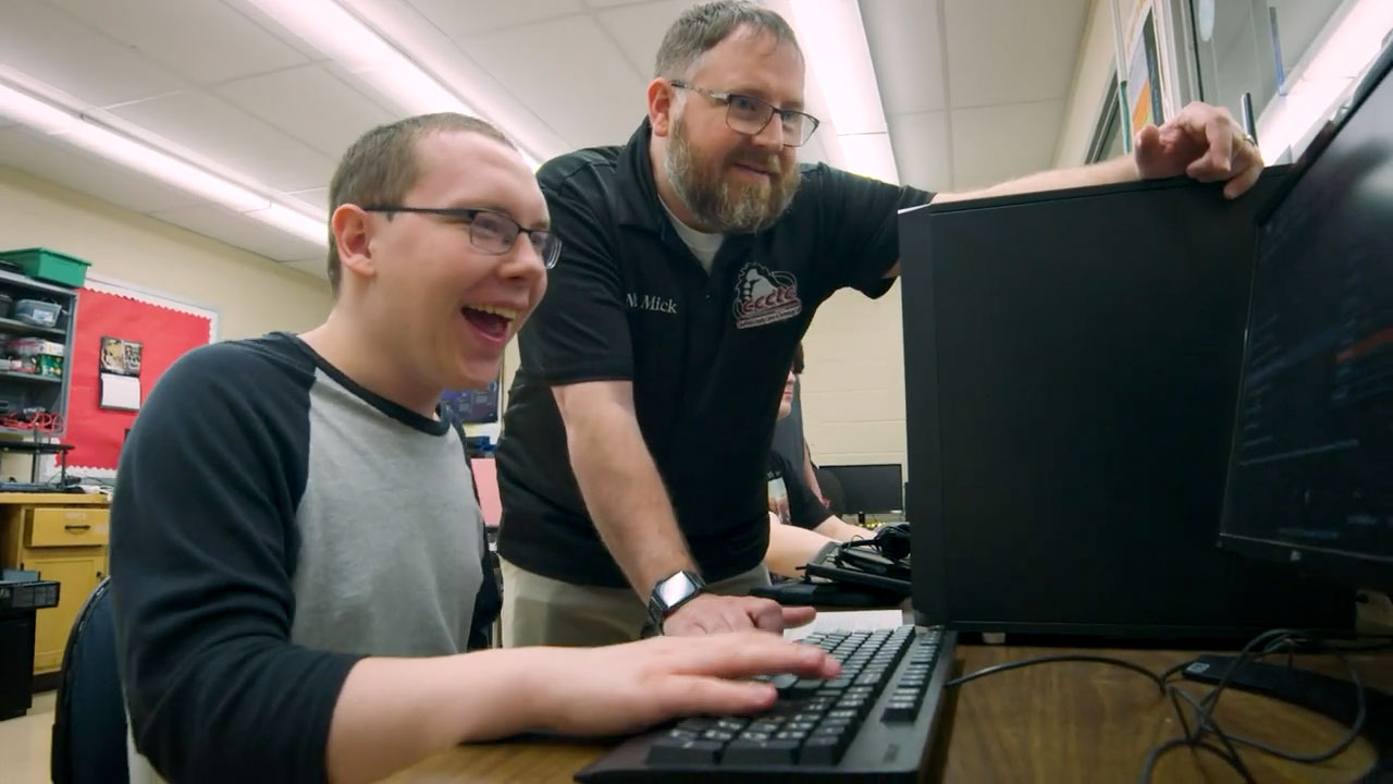 Teenage student using a desktop computer while bearded man in black shirt look over his shoulder at the screen.