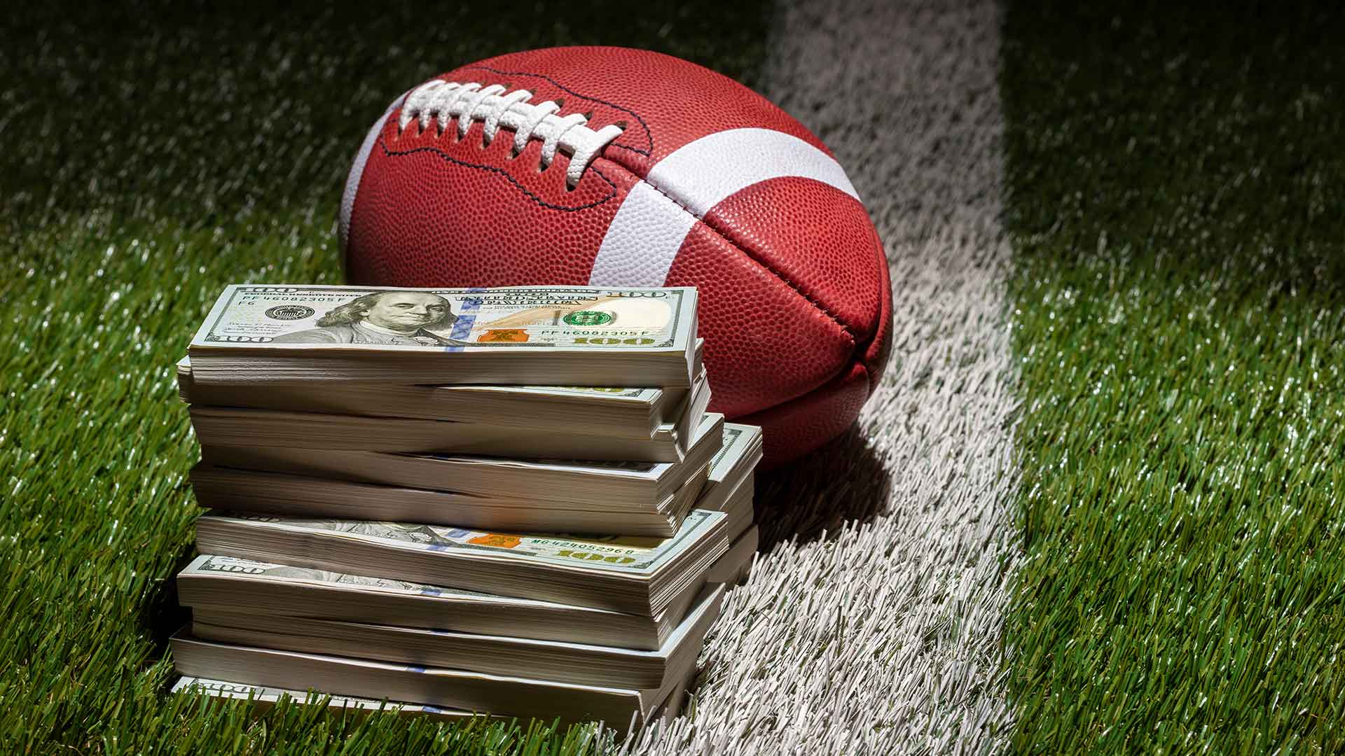 Football resting on field next to a large stack of 100 dollar bills.